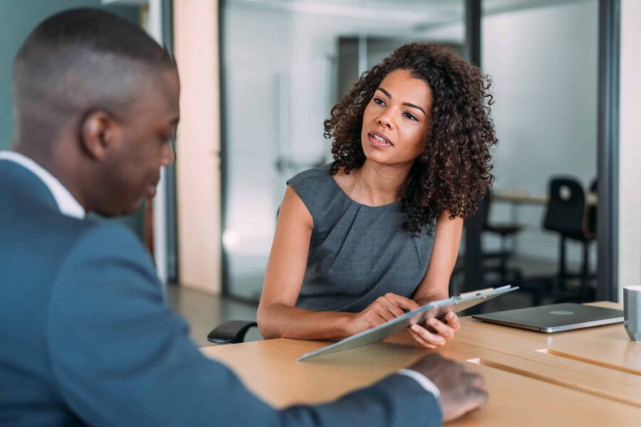 Two people in formal attire are having a discussion at a table in an office setting. One person is holding a tablet, presumably reviewing data collection results, while speaking. The other is listening attentively, likely focusing on the insights from recent market research.
